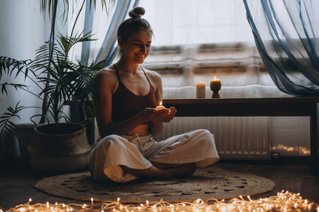 Young woman sitting on the floor, lights candles, enjoy meditation, do yoga exercise at home. Mental